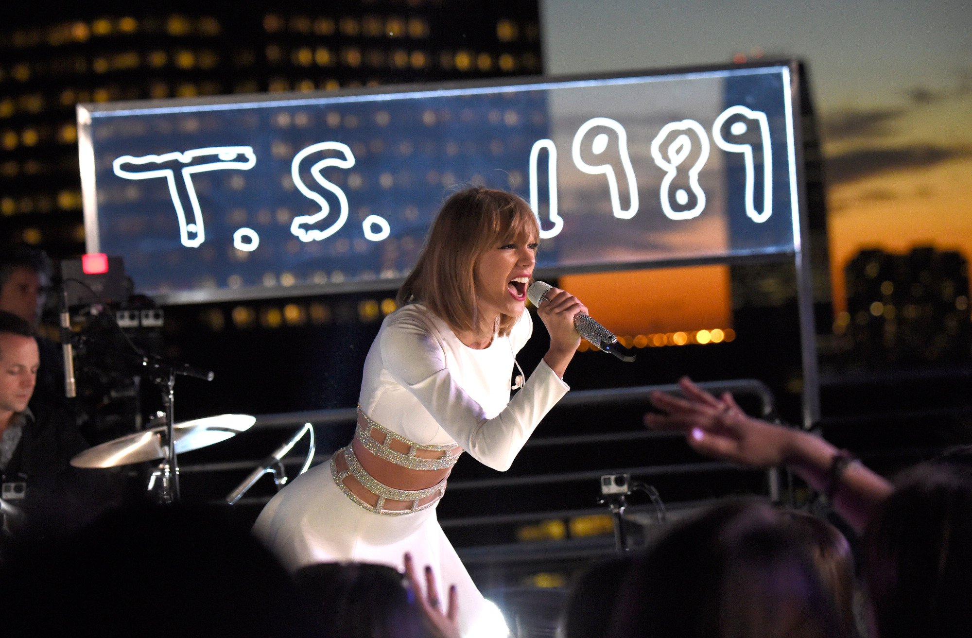 Taylor Swift performs during her 1989 Secret Session with iHeartRadio on October 27, 2014 in New York City. She is wearing white and a neon sign that says "T.S. 1989" glows behind her.