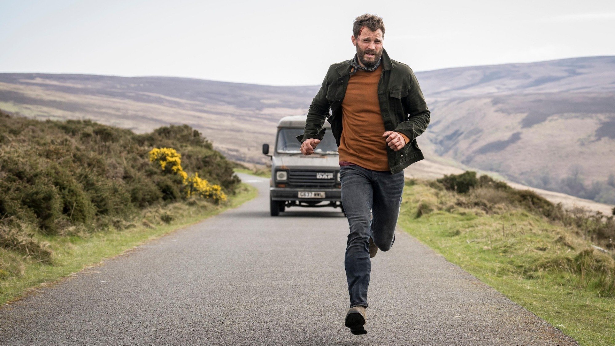 A man runs down a country road with a car in pursuit.