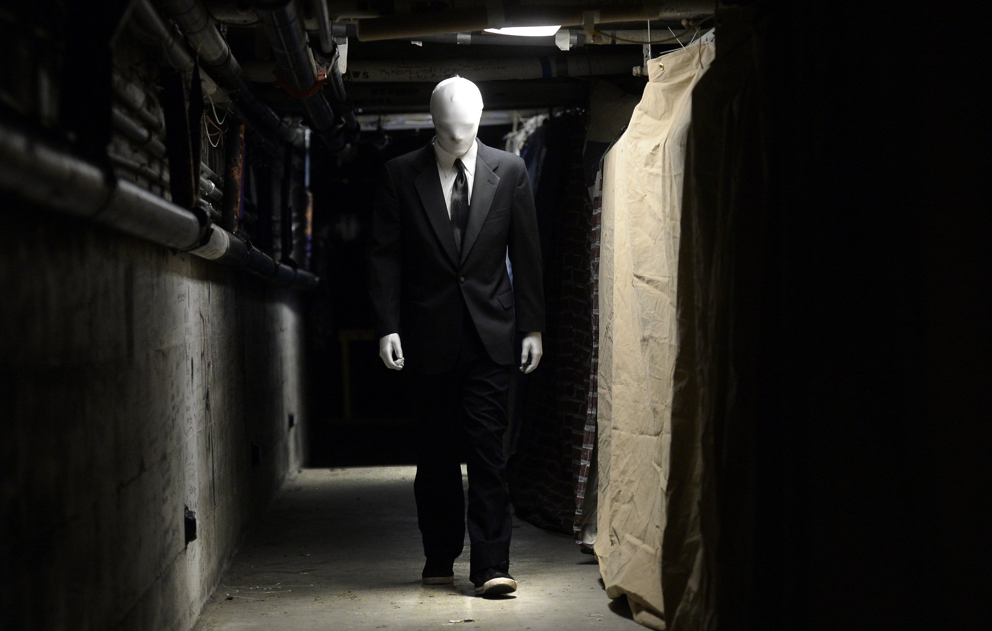 Someone dressed as Slender Man walks down a dimly lit hallway.