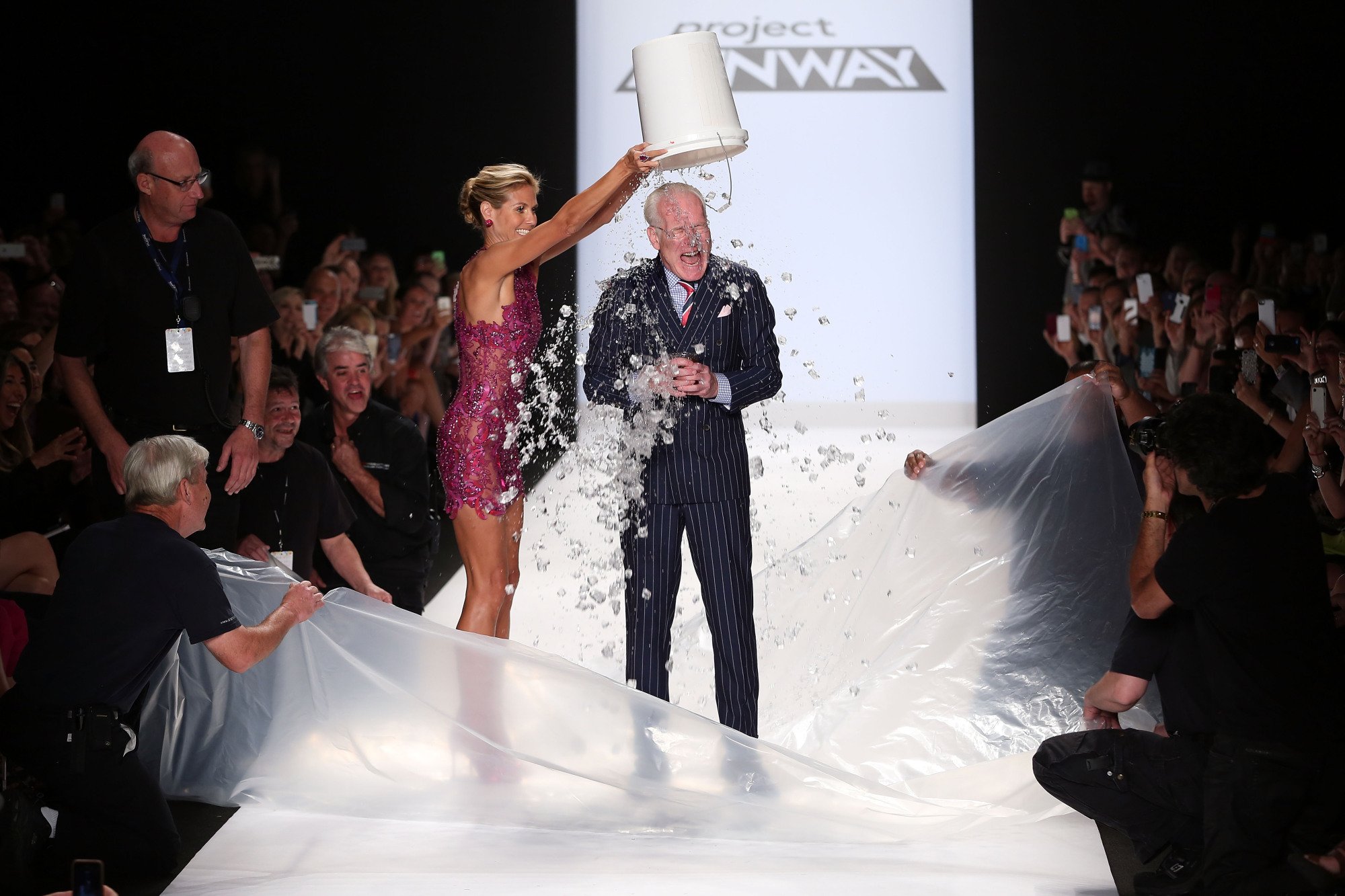 Heidi Klum (in a dark pink sequin dress) dumps a bucket of ice water on Tim Gunn (in a suit) at the Project Runway Season 13 Finale Show on September 5, 2014. They are on a white runway.