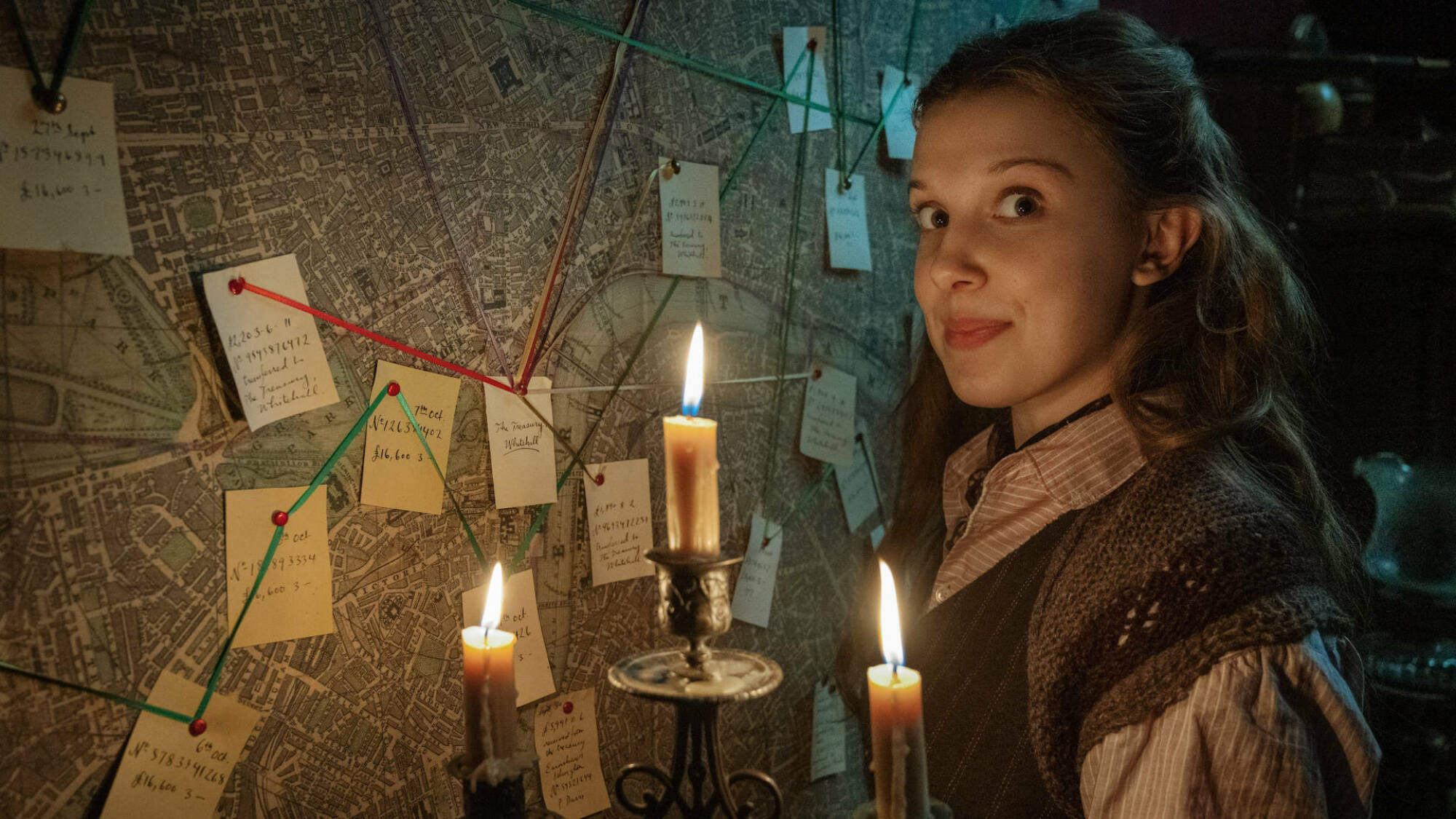 Millie Bobbie Brown stands by a red yarn wall with a candelabra in the film "Enola Holmes."