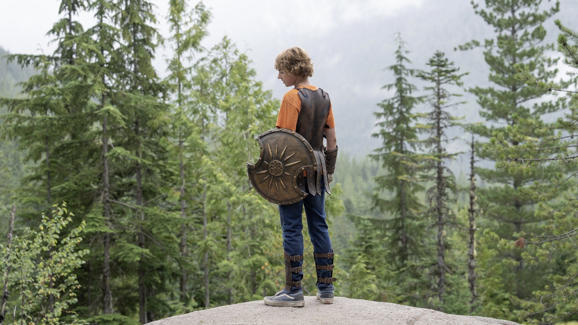 A young boy in Grecian armor and an orange T-shirt stands on top of a hill overlooking a forest.