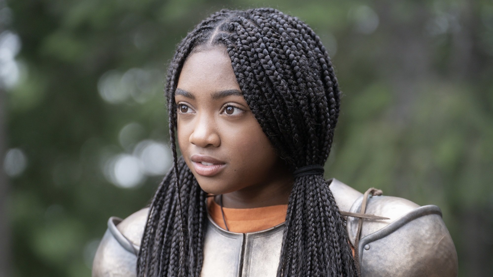 A girl with braids wearing Grecian armor over an orange T-shirt.