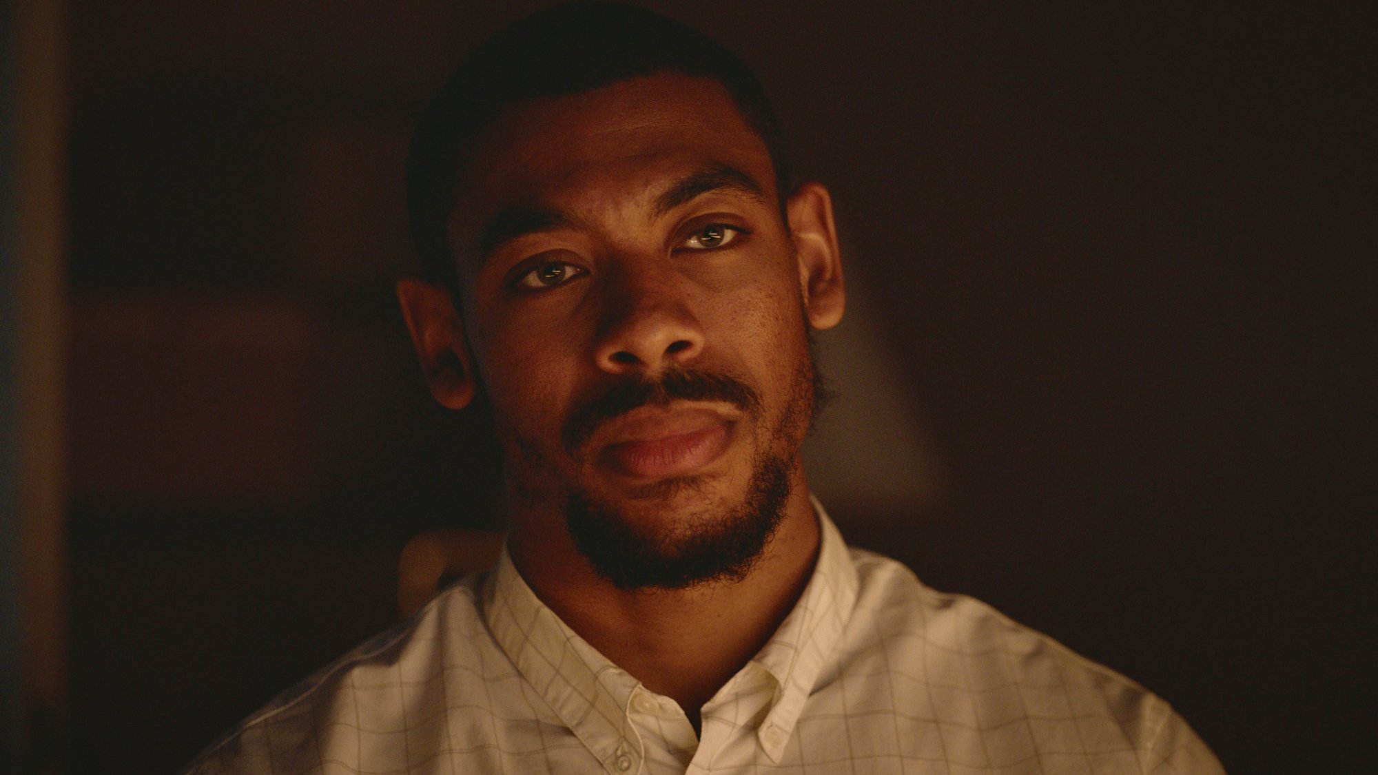 A man wearing a white shirt stands looking pensive in a low-lit room.