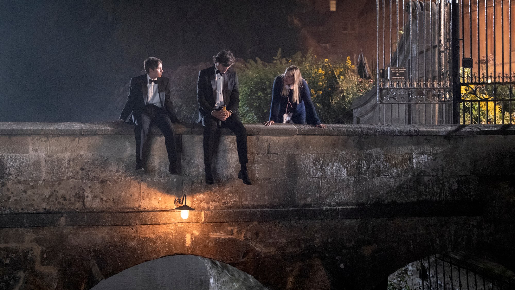 Two young men in tuxedos sit on a stone bridge.