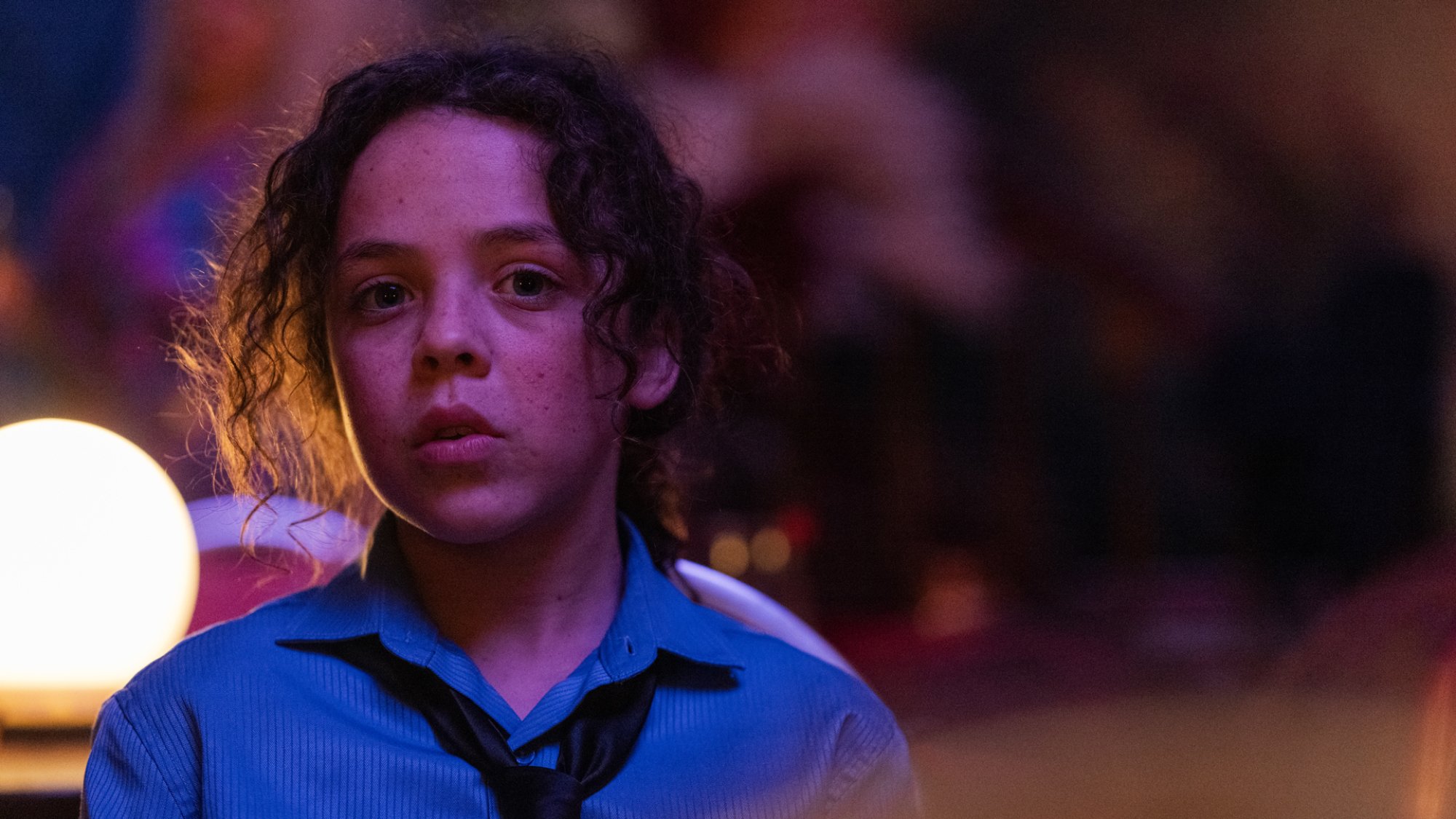 A young boy in a shirt and tie stands in a purple light.