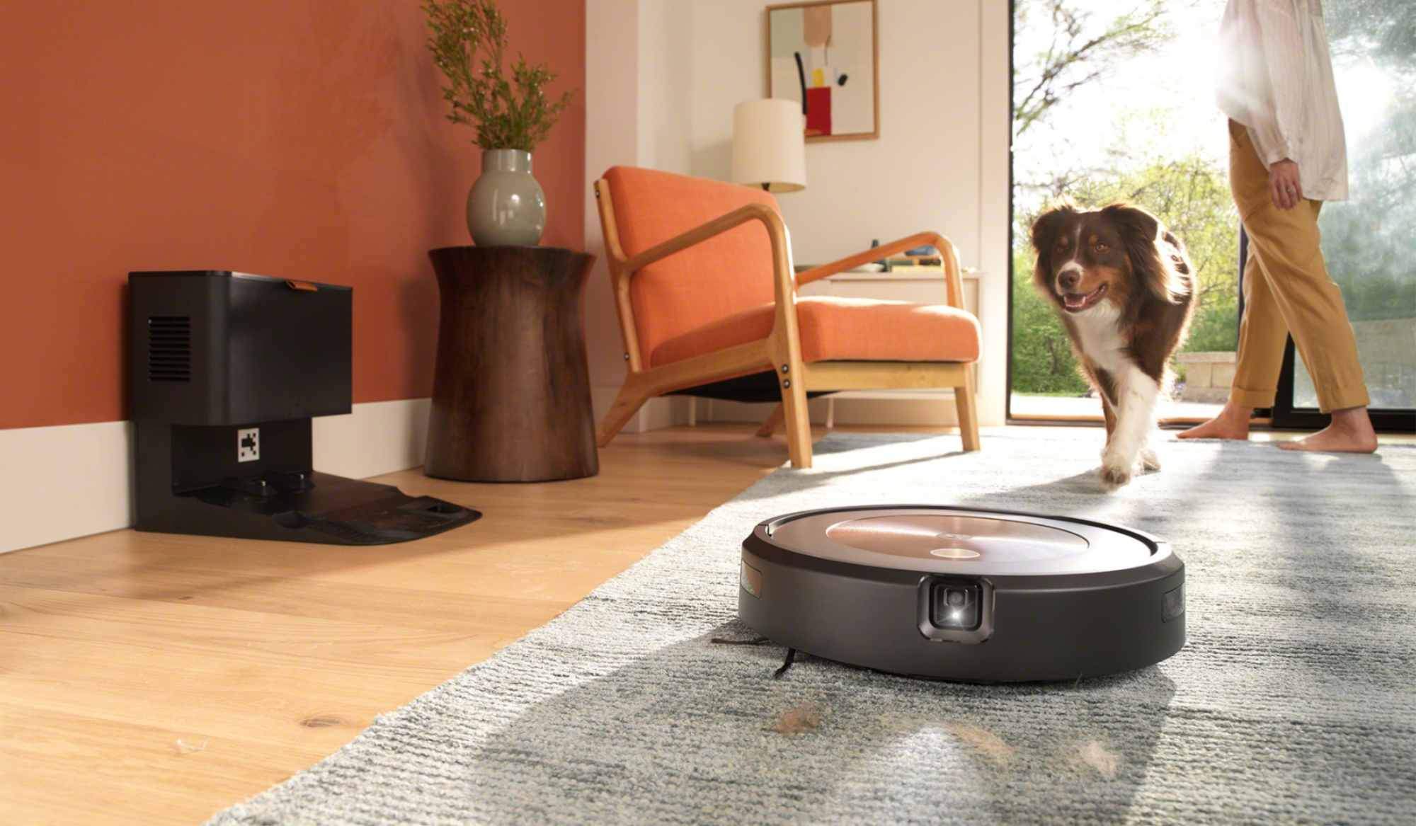 Roomba with camera and headlight cleaning rug with dock, dog, and person in background