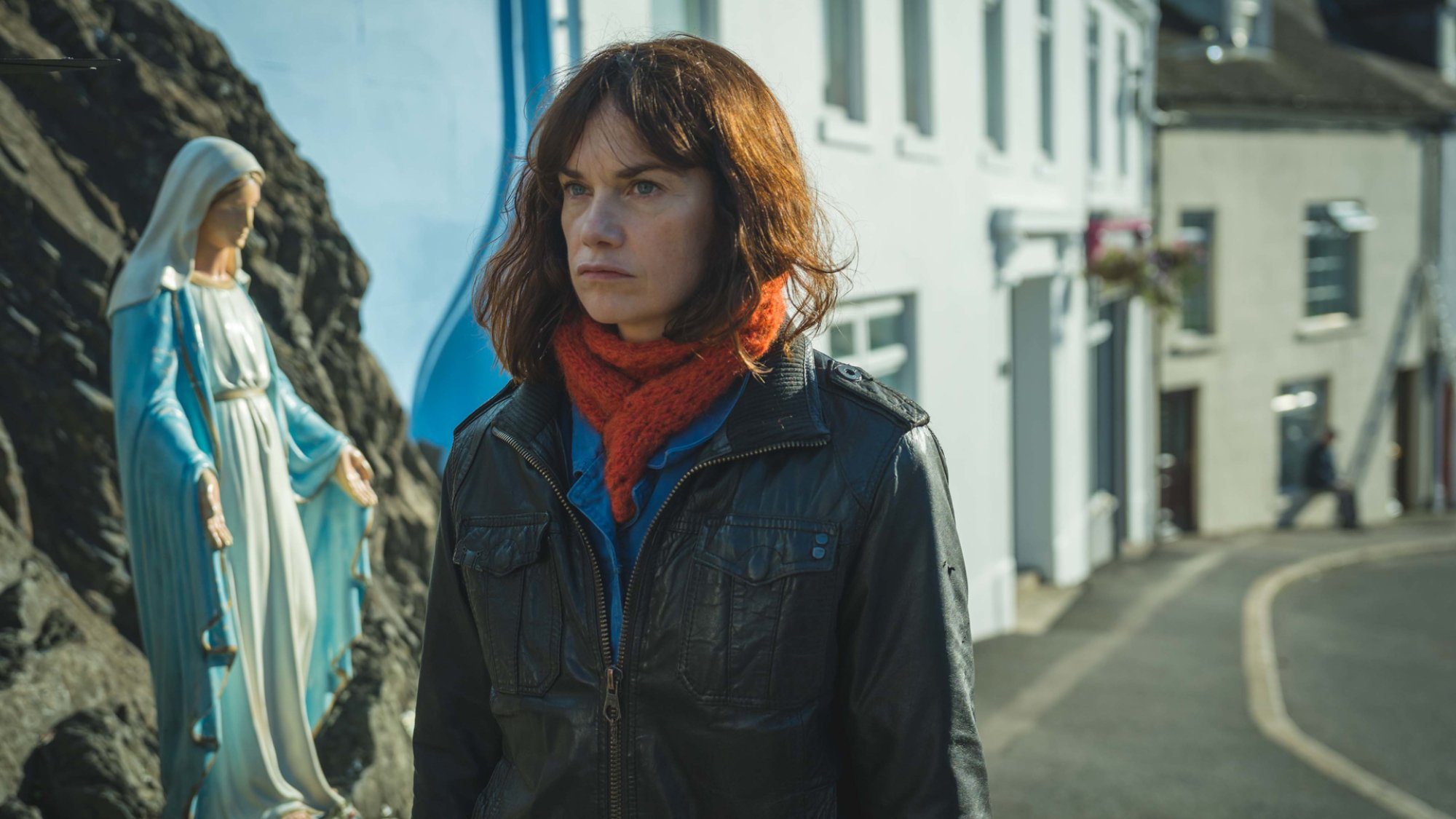 Ruth Wilson walks down a street in a small town in Ireland, passing a religious figure of the Madonna.