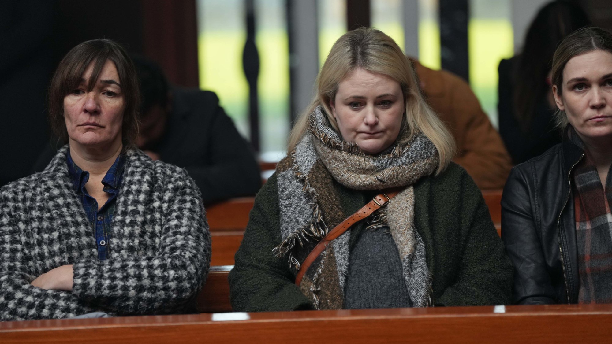 Three women looking glum in a church pew, wearing dull-coloured warm clothing.