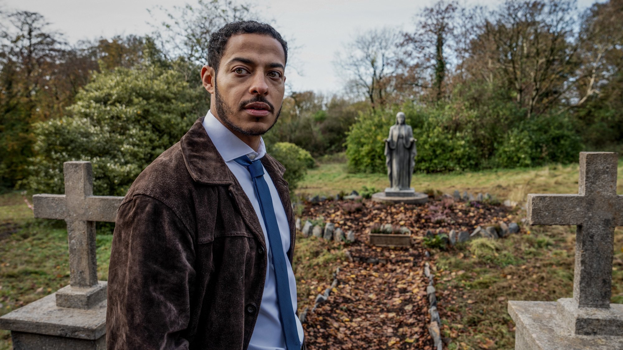 Daryl McCormack stands in a graveyard investigating something in the TV show "The Woman in the Wall"