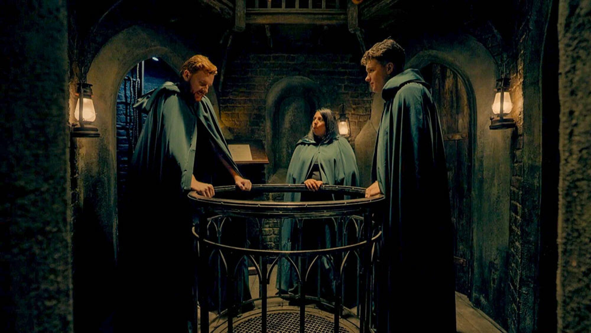 Two men and a woman stand in a dark room wearing black cloaks.