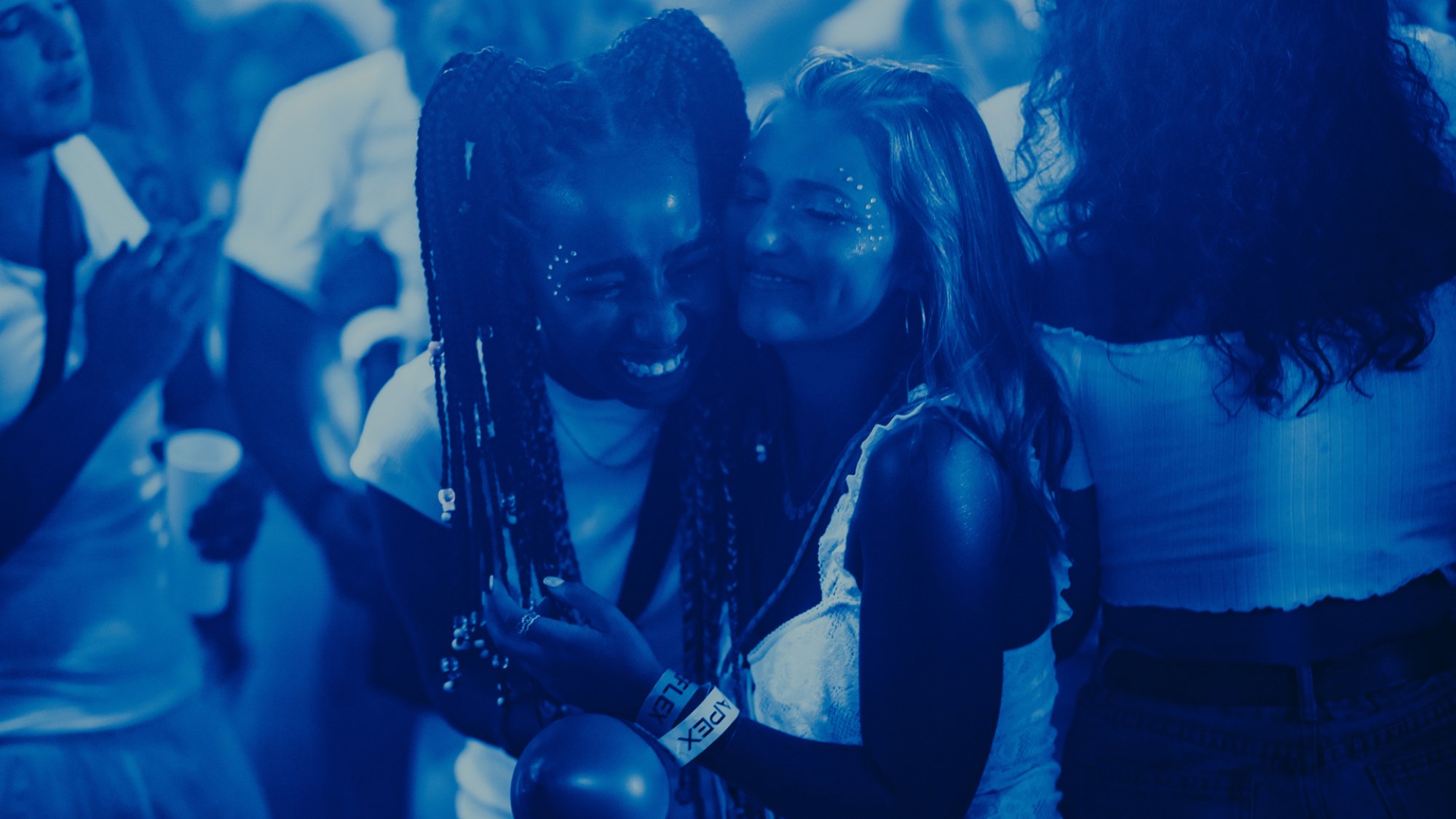 Two teen girls hug on a dancefloor with their eyes closed and smiling.