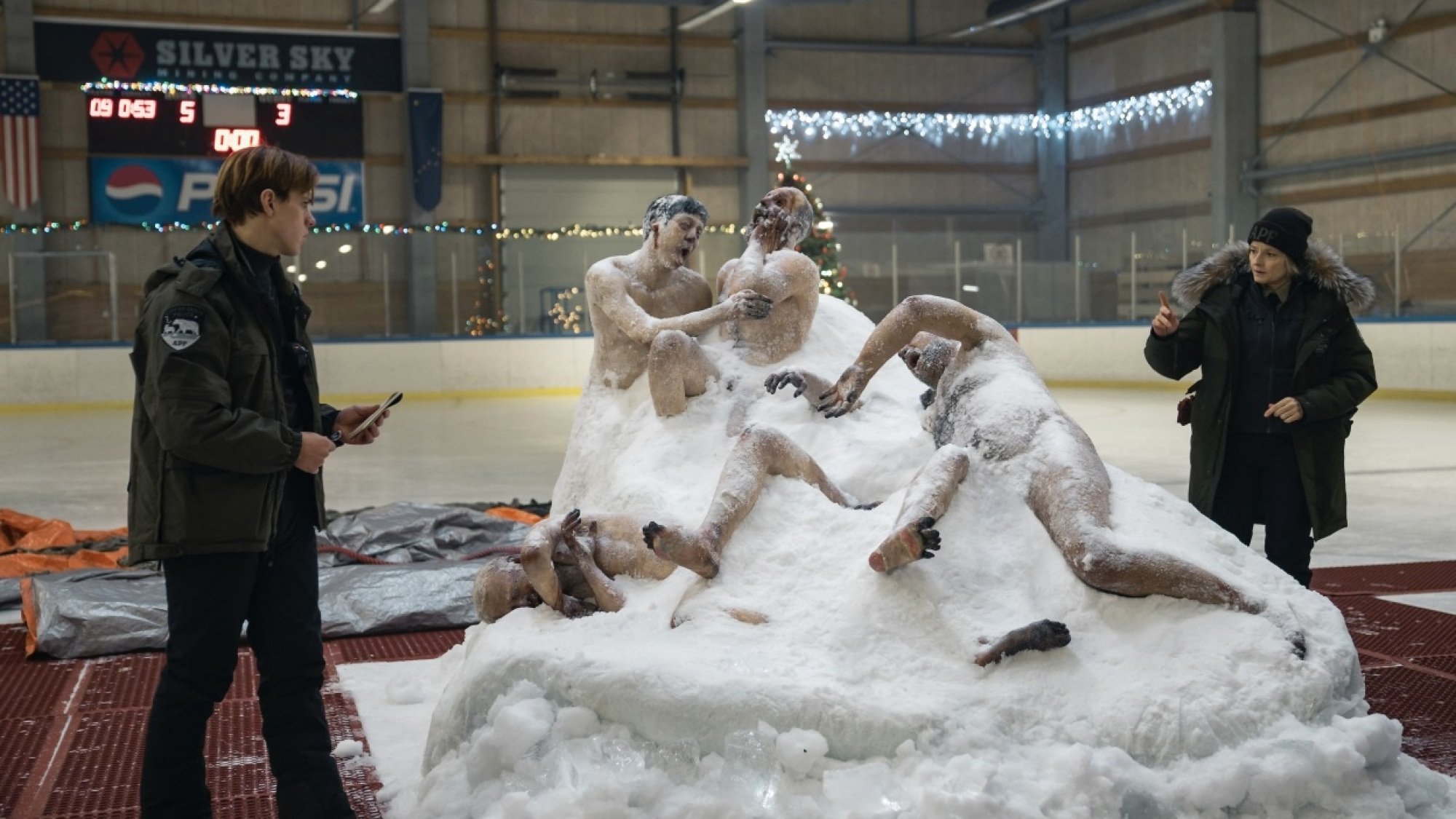 A man and woman in police uniforms inspect a frozen clump of corpses thawing on an ice rink.