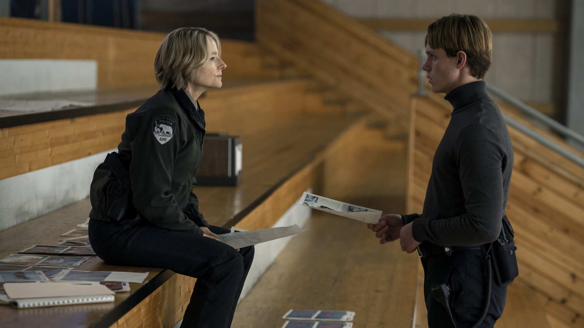 A detective and her protege sit in a stadium piecing together clues on printed paper.