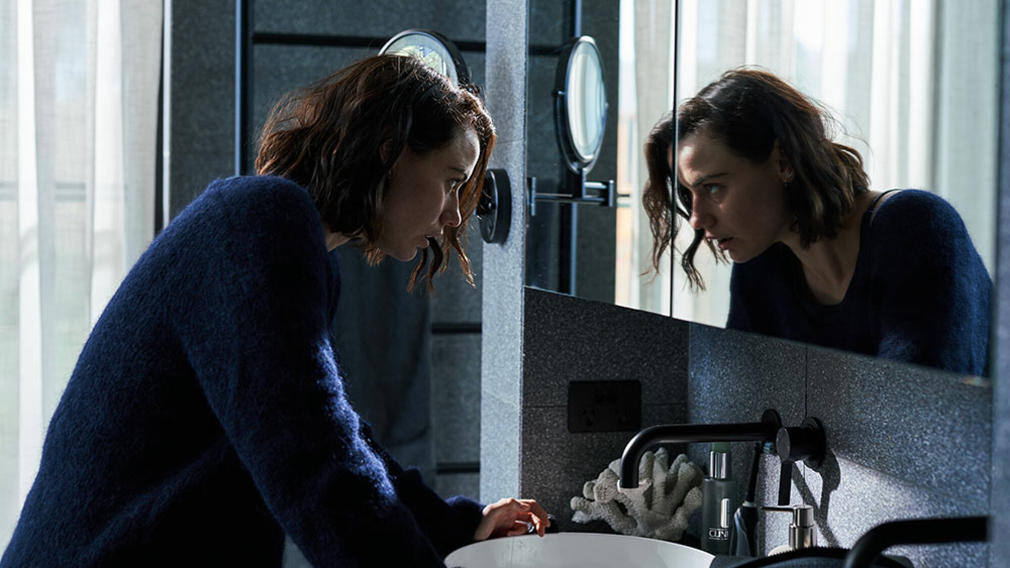 A woman stands in front of a bathroom mirror, staring at her reflection.