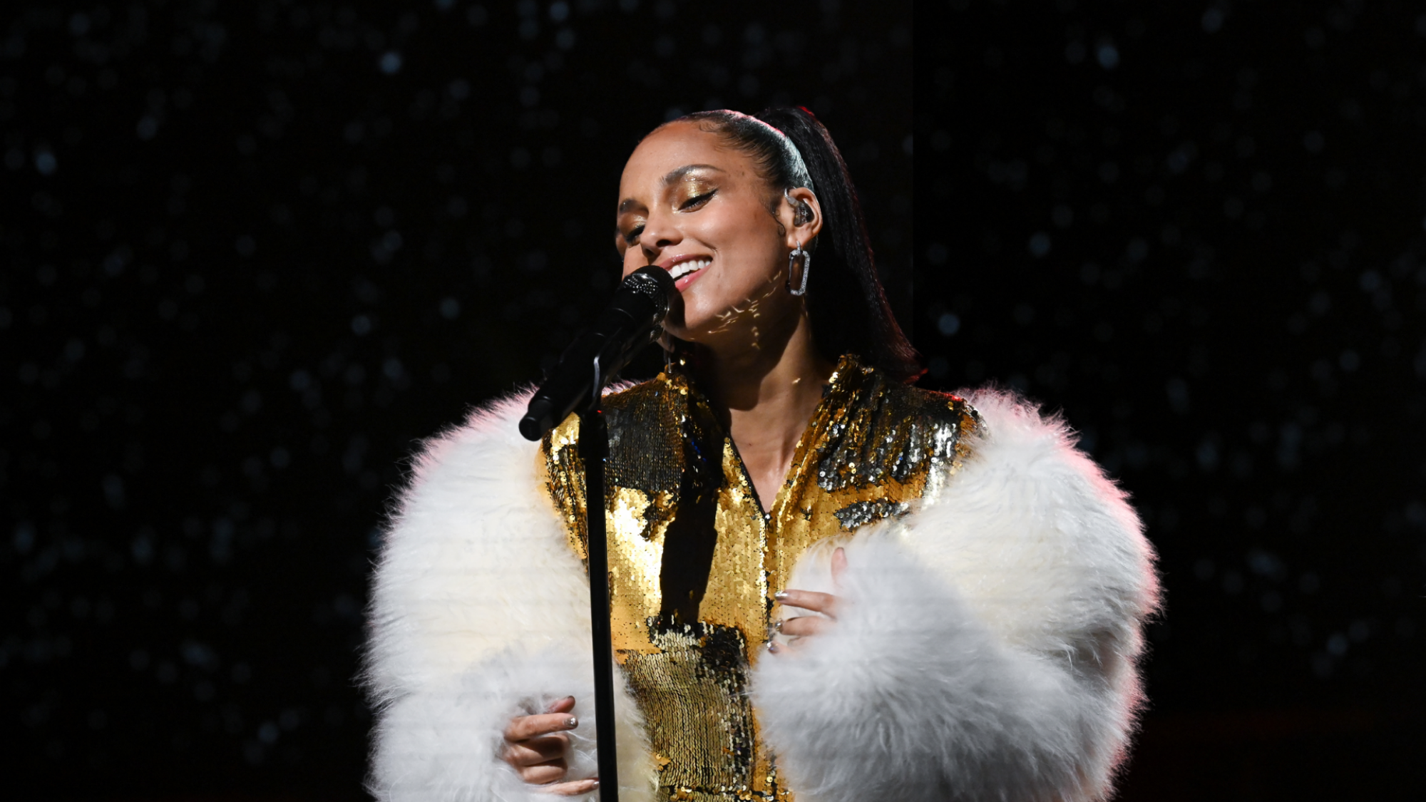 Alicia Keys sings into a black microphone on a stand. She is wearing a gold dress and a white fur stole.