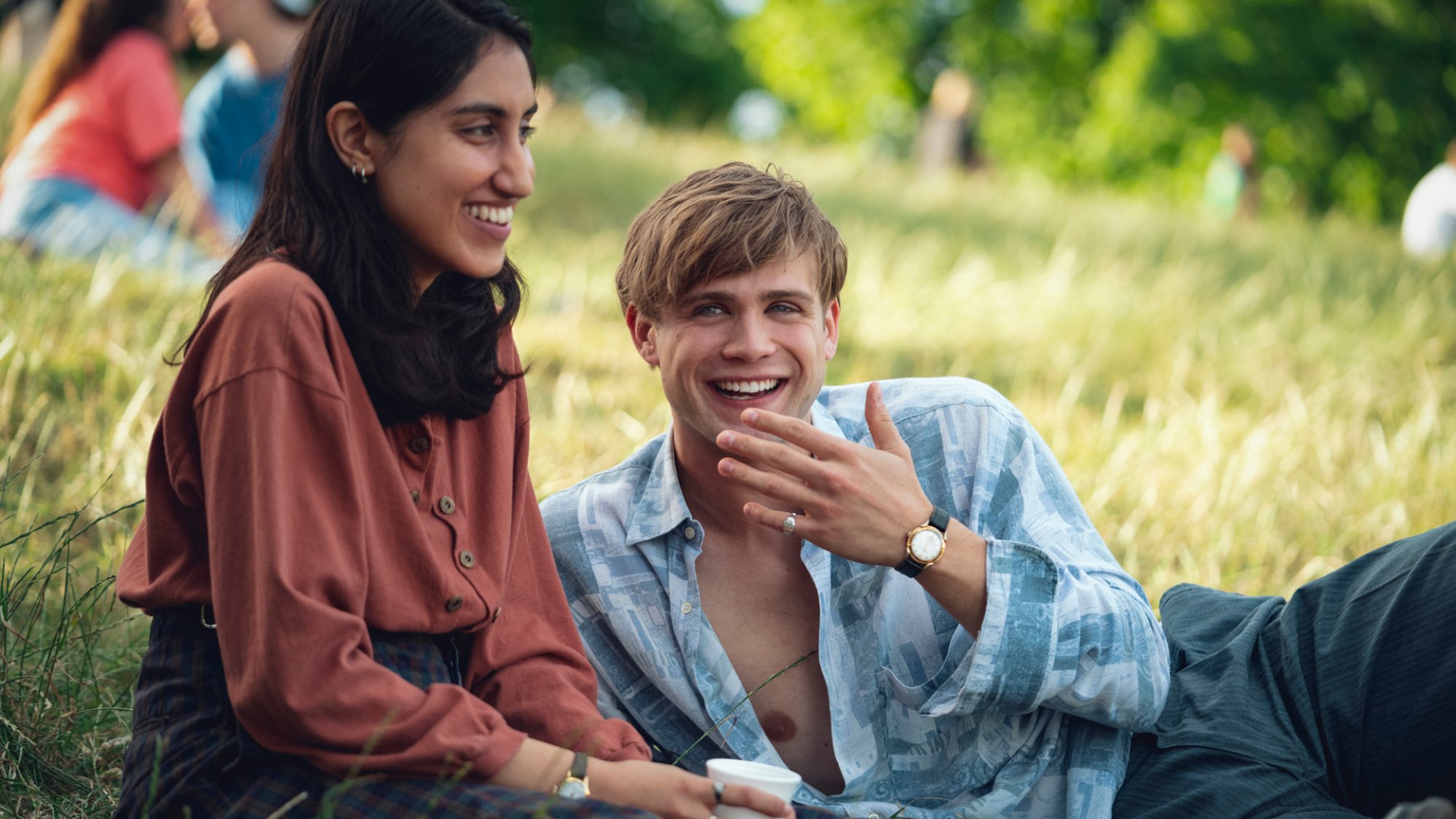 Two young people sit on a grassy hill in the sun laughing. 