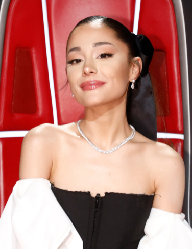 Ariana Grande in her The Voice chair with dark eyebrows and hair, with a red lip.