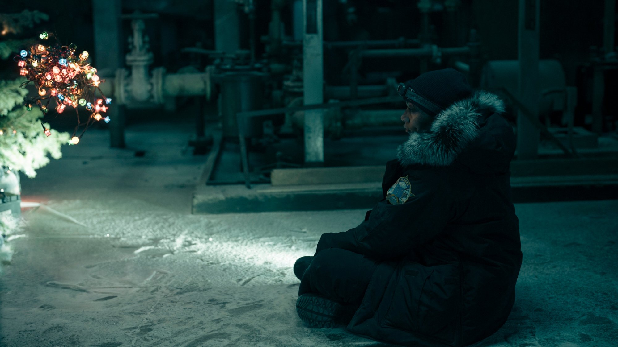 Kali Reis sits by a Christmas tree in a dark dilapidated room.