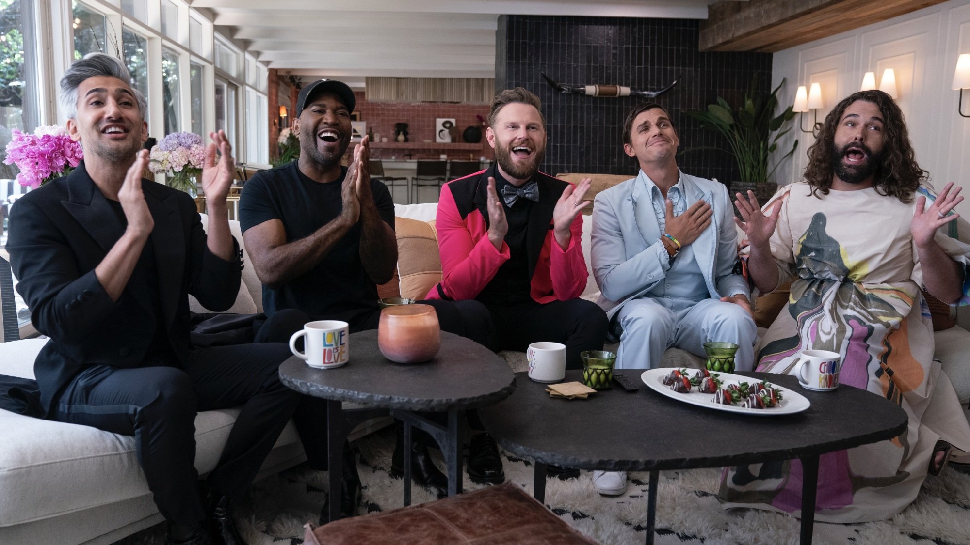 Four men and one nonbinary person, all in fancy clothing, sit on a couch and applaud.