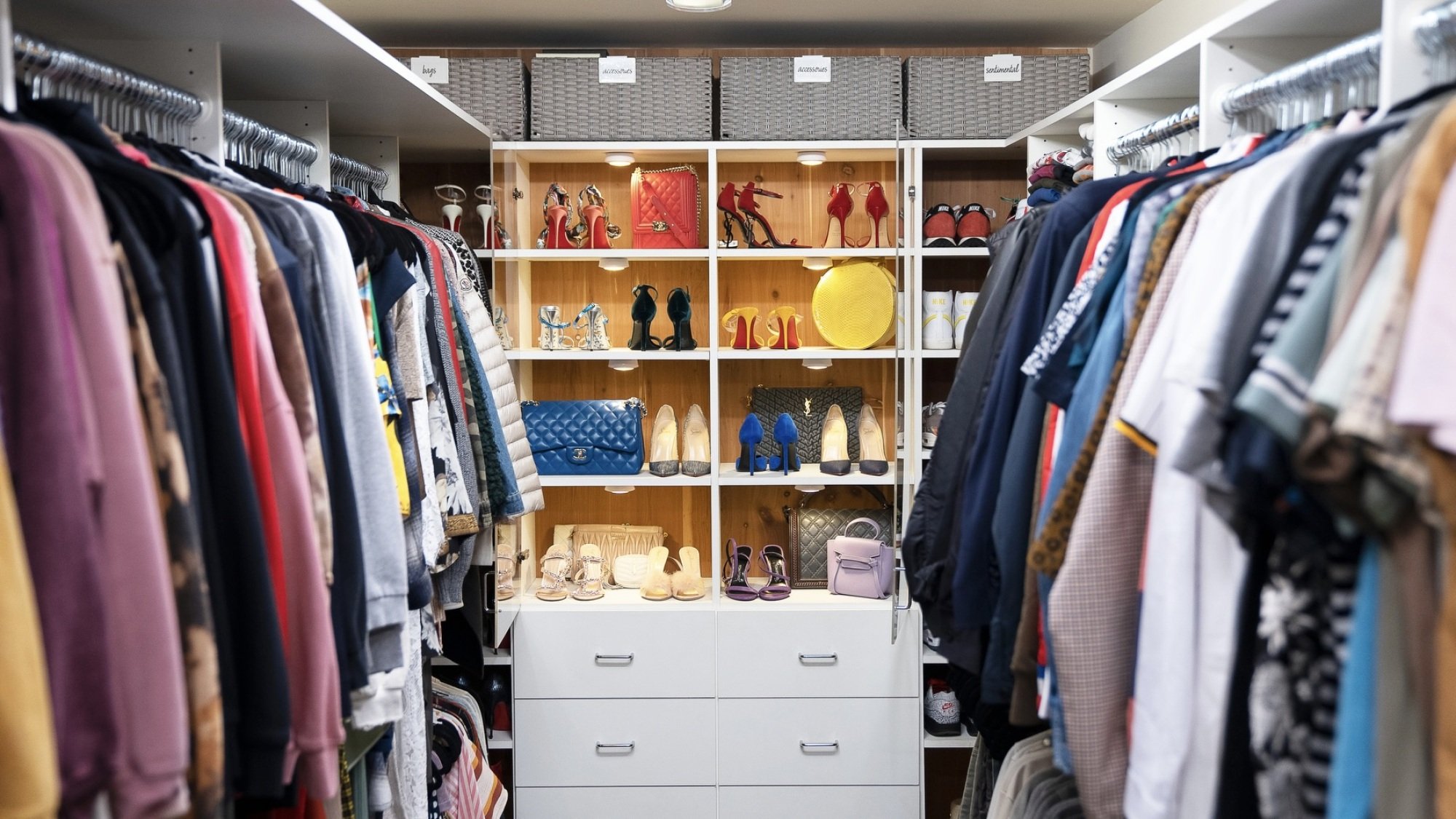 A very organized walk-in closet, with clothes on hangers on two different walls and shoes and purses on the far end of the room.