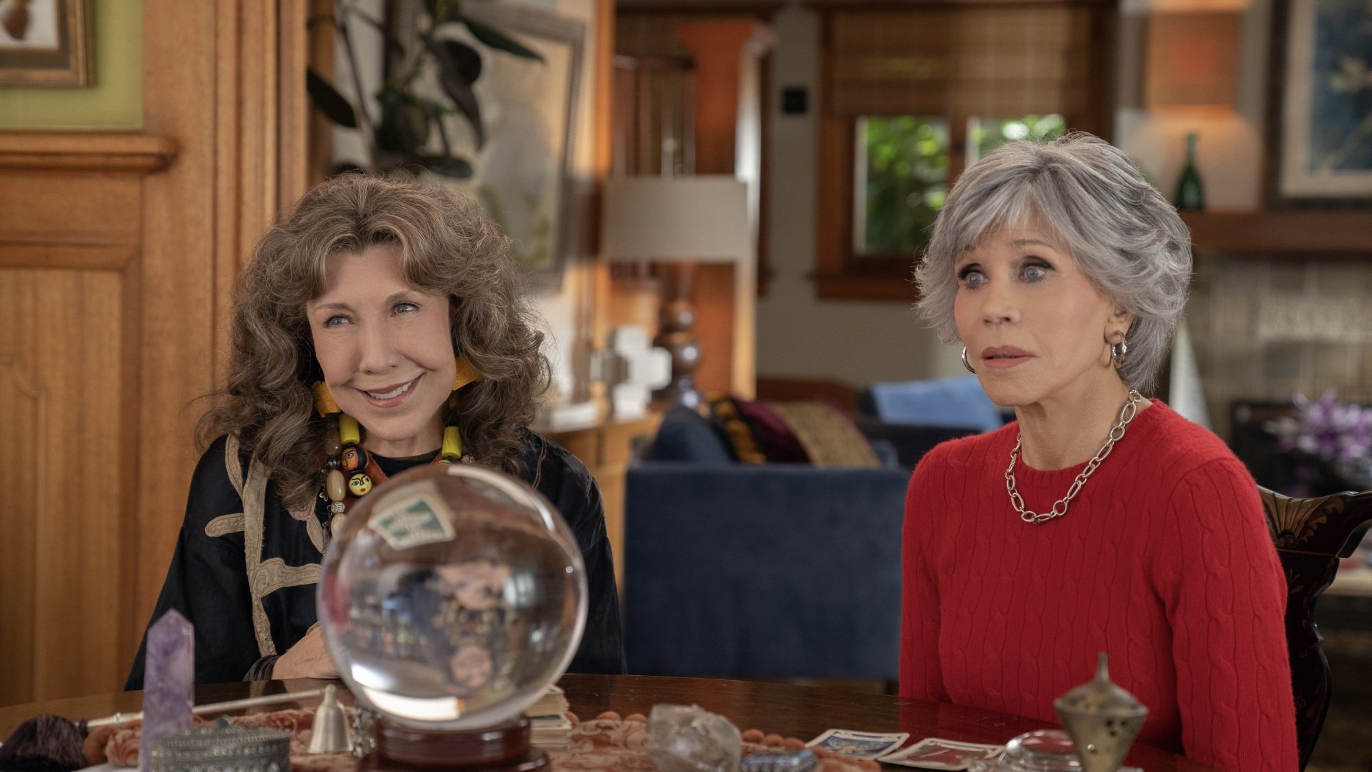 Two older women, one in a red sweater, one in a black draped shirt, sit at a table with a crystal ball on it.