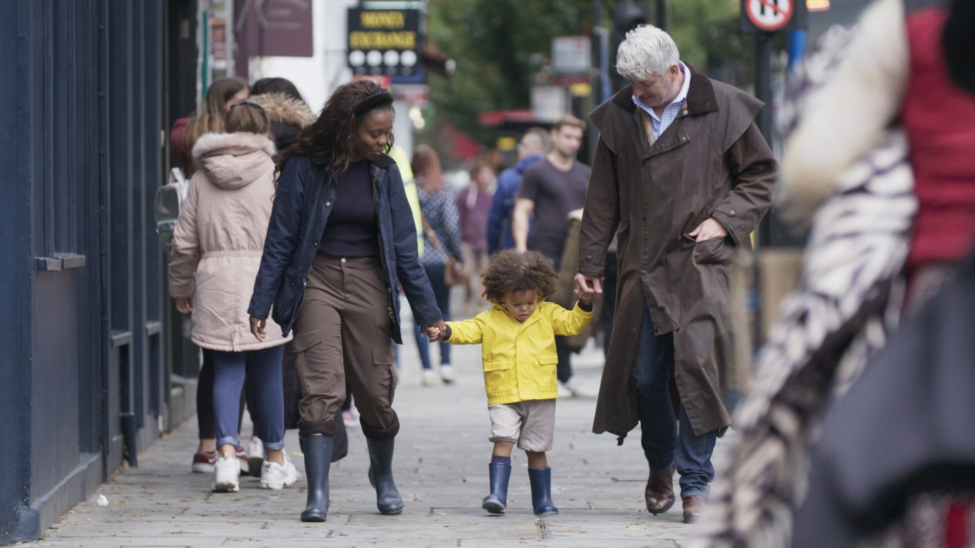 A man and woman hold the hands of a small child walking between them on the street. The child is wearing a yellow raincoat.