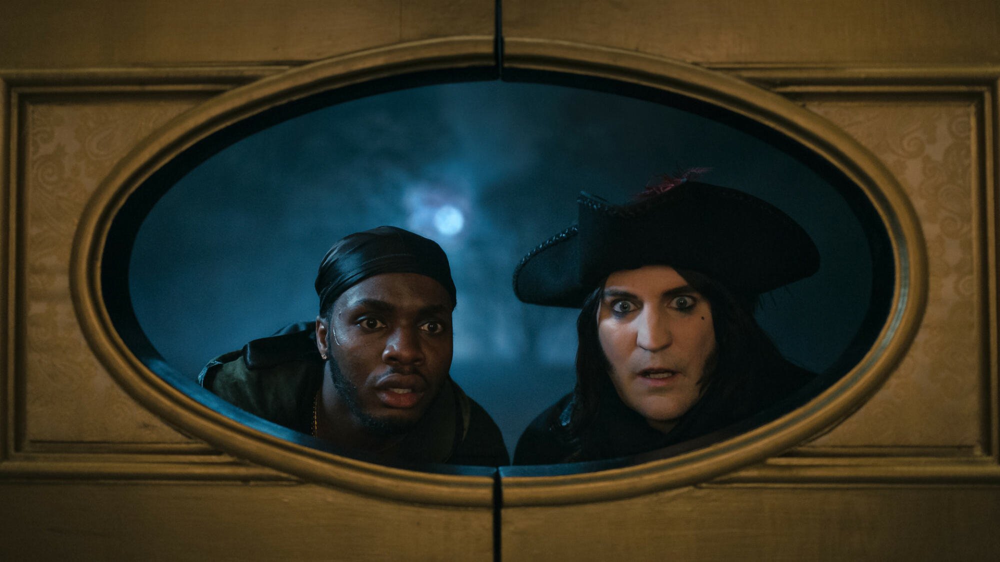 Duayne Boachie and Noel Fielding in 18th century garb peer through a round window.
