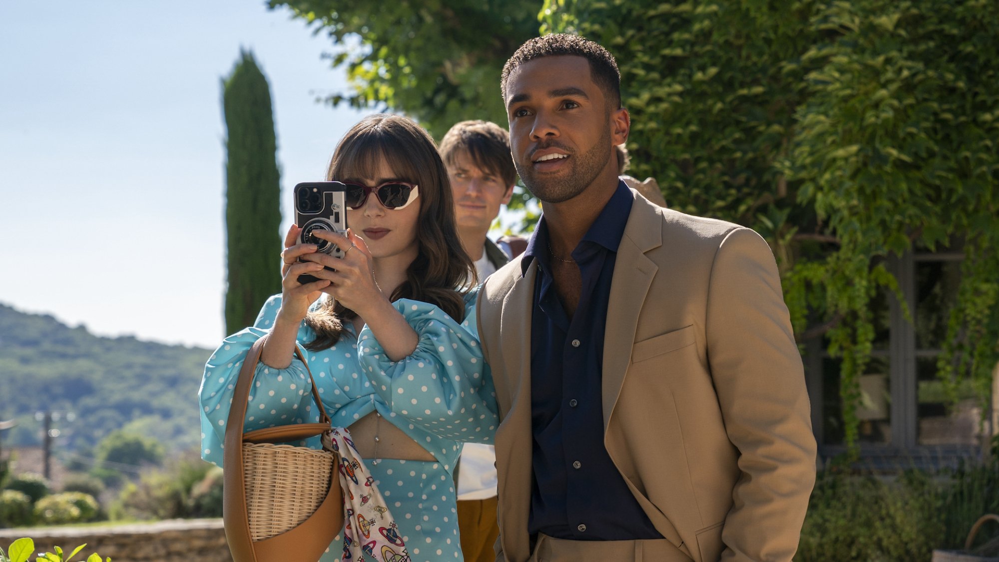 A well-dressed couple stand looking at a view in an estate's leafy grounds, taking a photo on a phone.