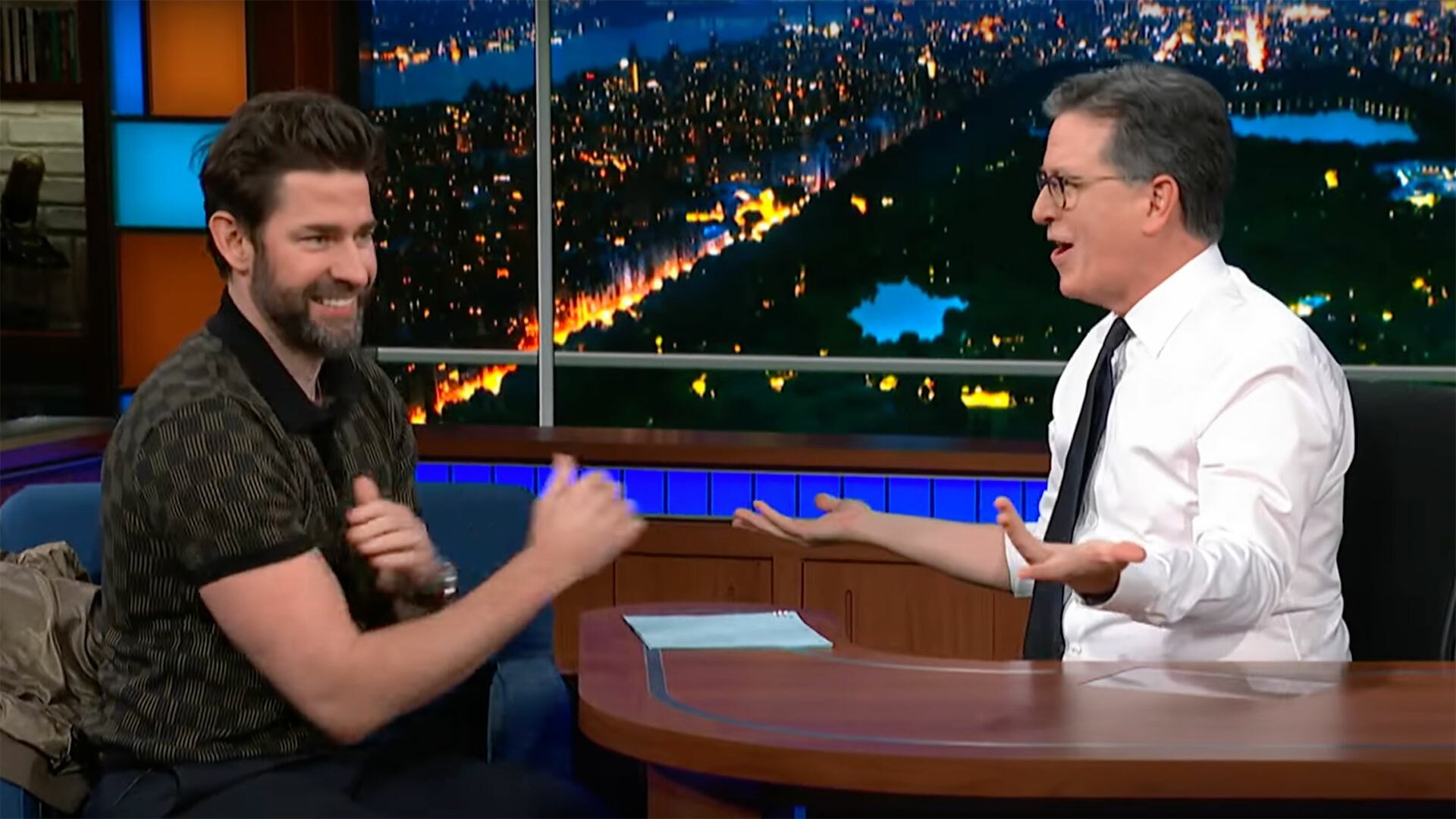 Two men face each other, laughing, over a talk show desk.