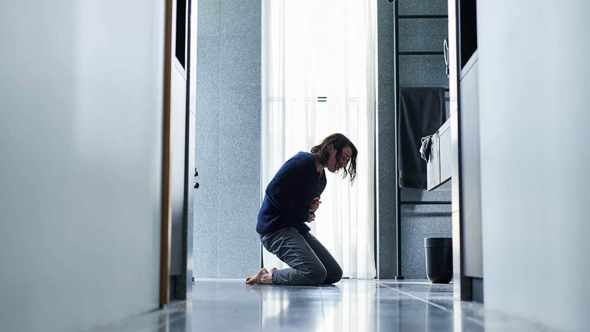 A woman kneels on the floor of a bathroom, clutching her stomach.