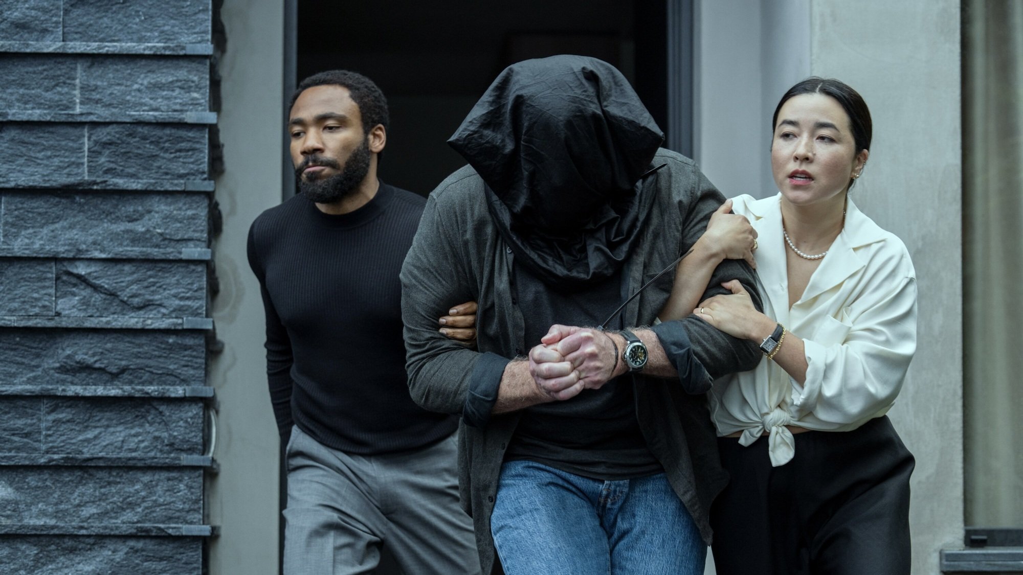 A man and woman escort a man with a black bag over his head out of a house.