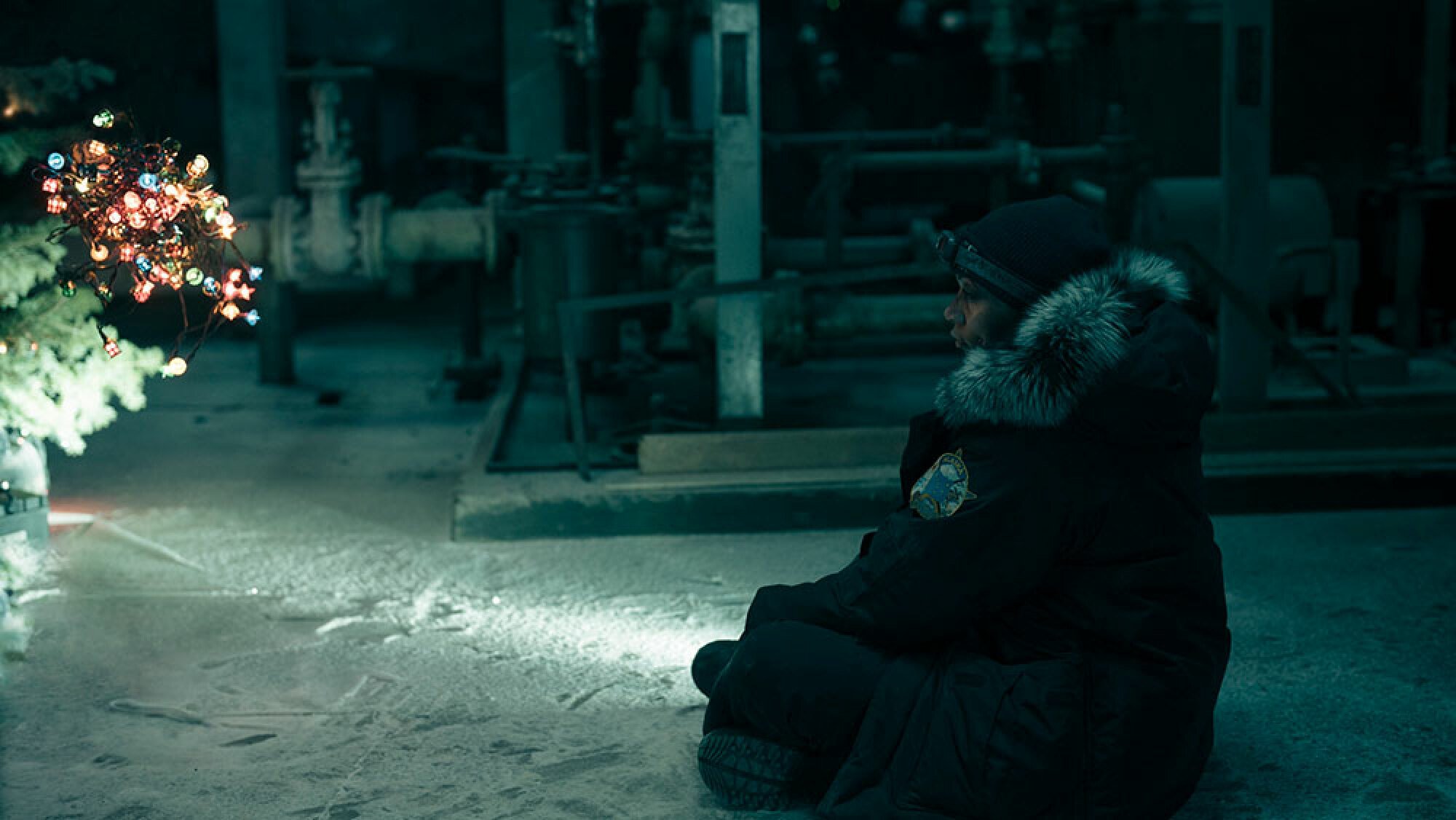 A woman sits on the cold ground, shining her torch into the darkness at a Christmas tree.