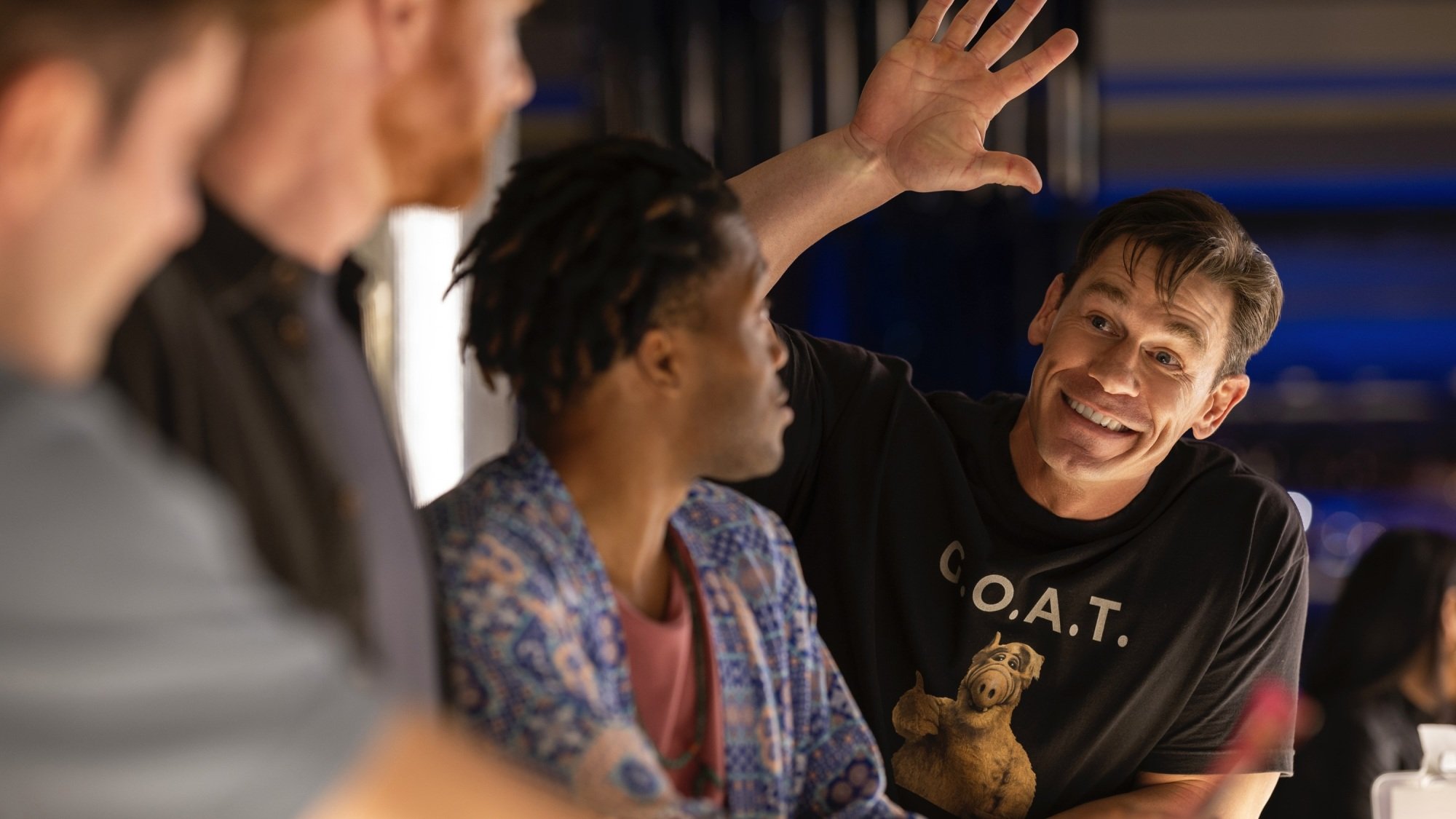 A man in an ALF shirt waves at three other men across the bar.
