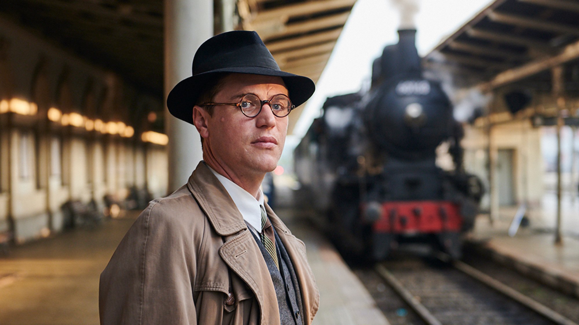 A young man wearing glasses stands on a train platform with a steam train in the background.