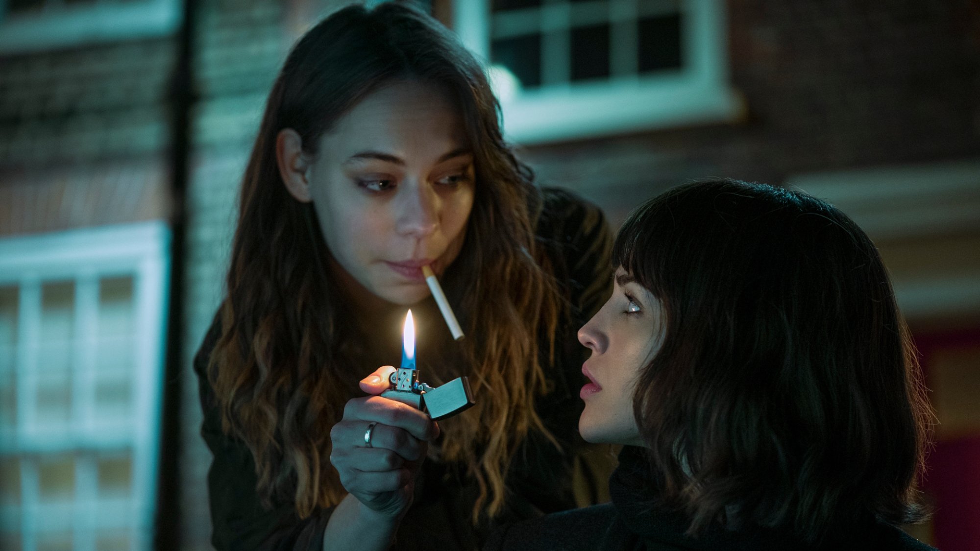 A woman with a cigarette in her mouth holds a lighter aflame for another woman.