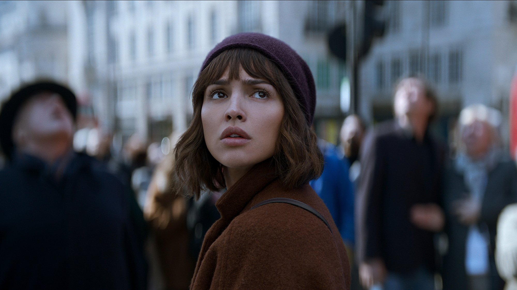 A woman stands in the city of London staring at the sky looking worried.