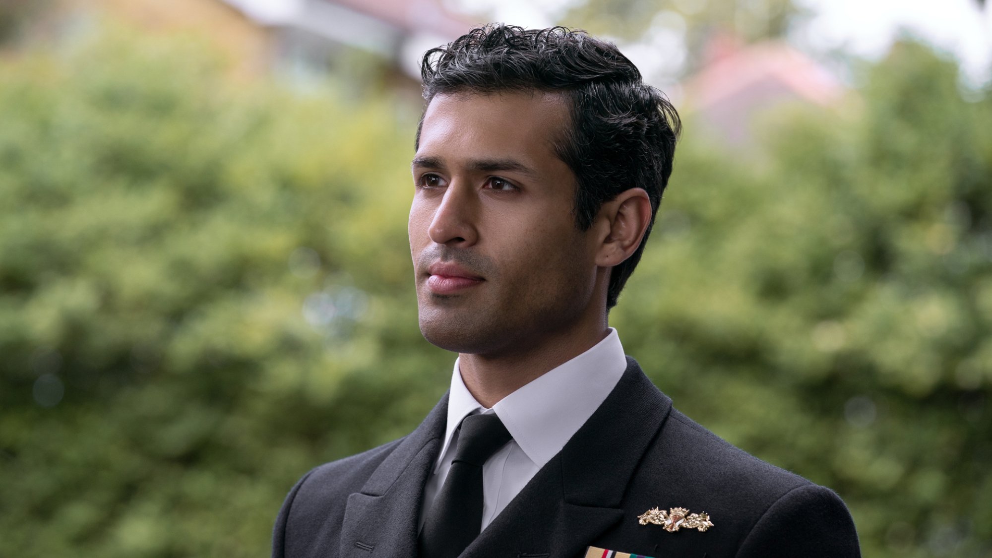 A military officer in a suit stands in front of a green hedge.