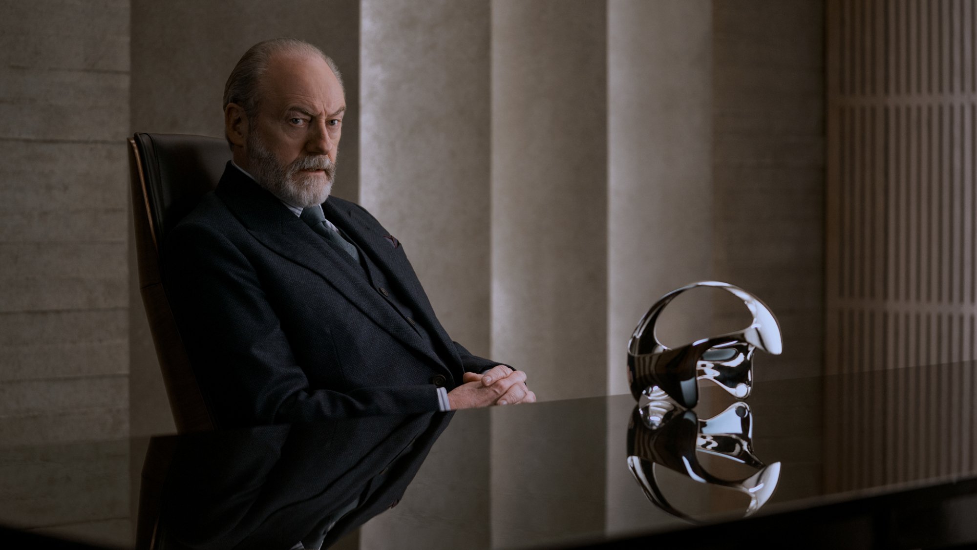 A man in a suit sits behind a polished black desk, on which sits a silver VR headset.