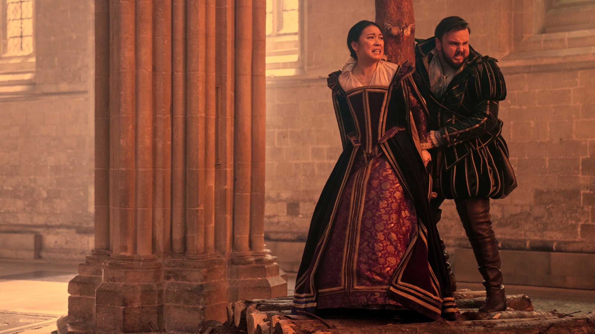Two characters in 15th century English costumes look frantic as the room they're in appears glowing.