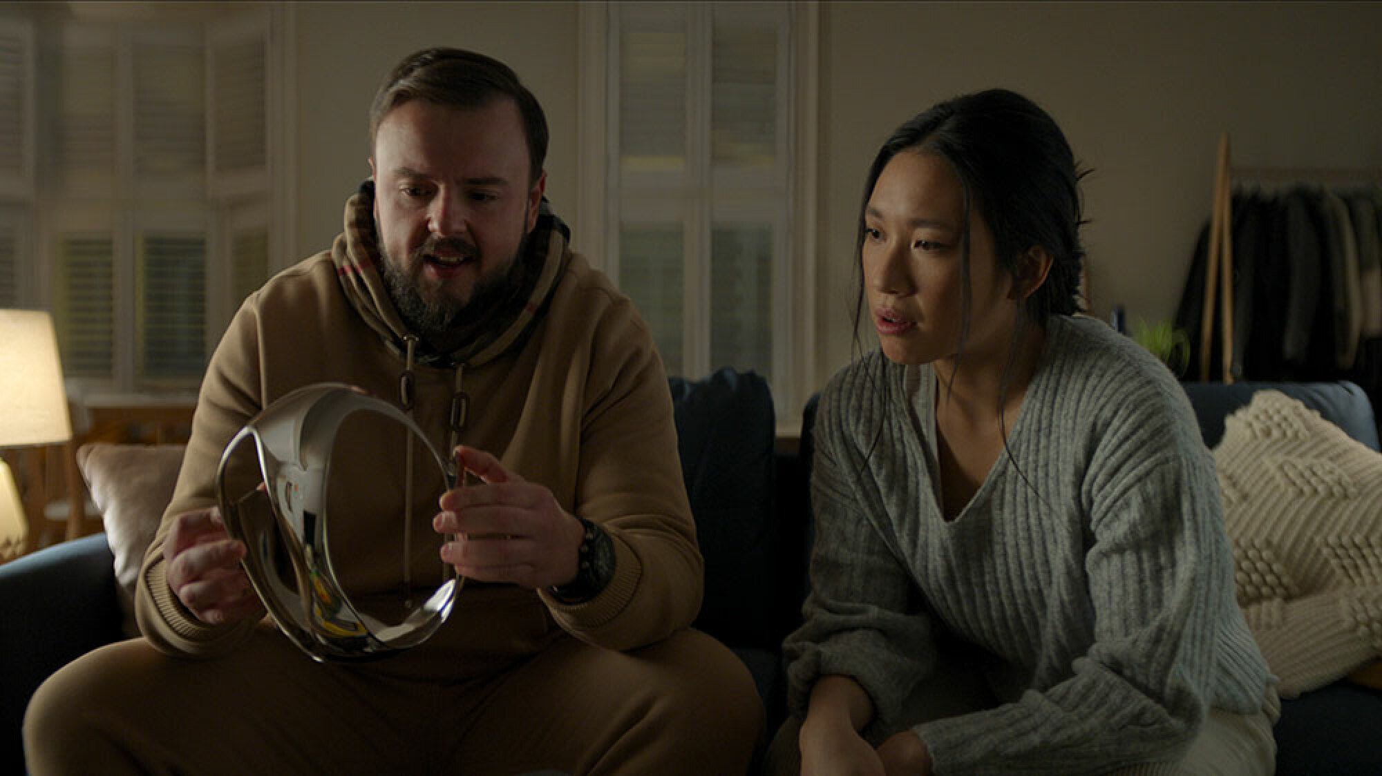 A man and a woman hold what looks like a shiny gold VR headset while sitting on a sofa.