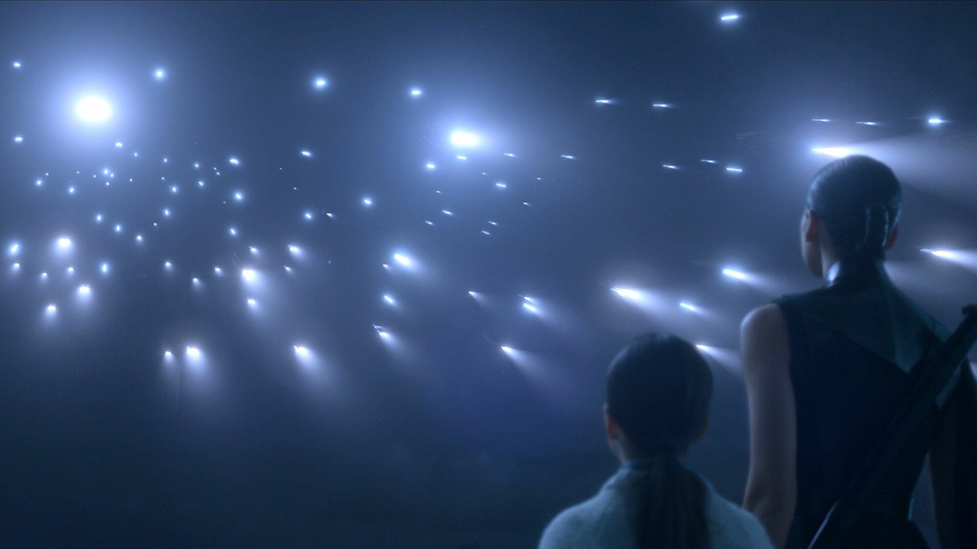 A woman with a sword on her back stands next to a little girl, looking up at bright lights in a dark sky.