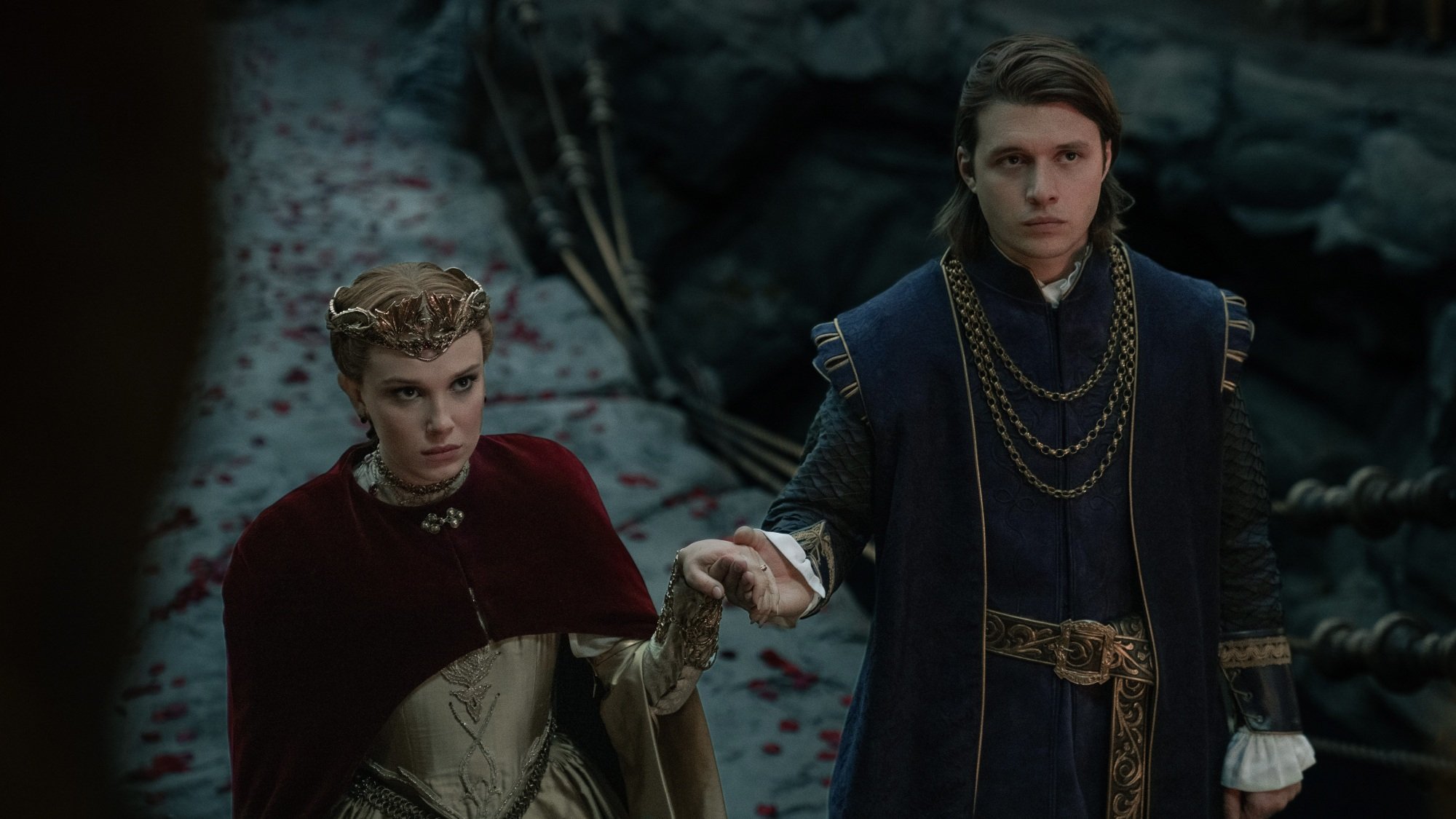 A woman in a red and gold gown and a man in a blue doublet hold hands.