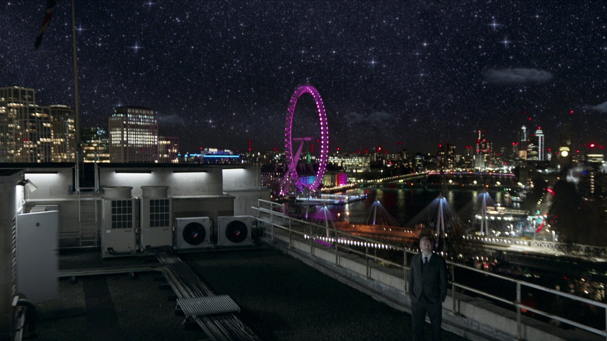 A man stands on a rooftop under the stars with the London skyline in the background.