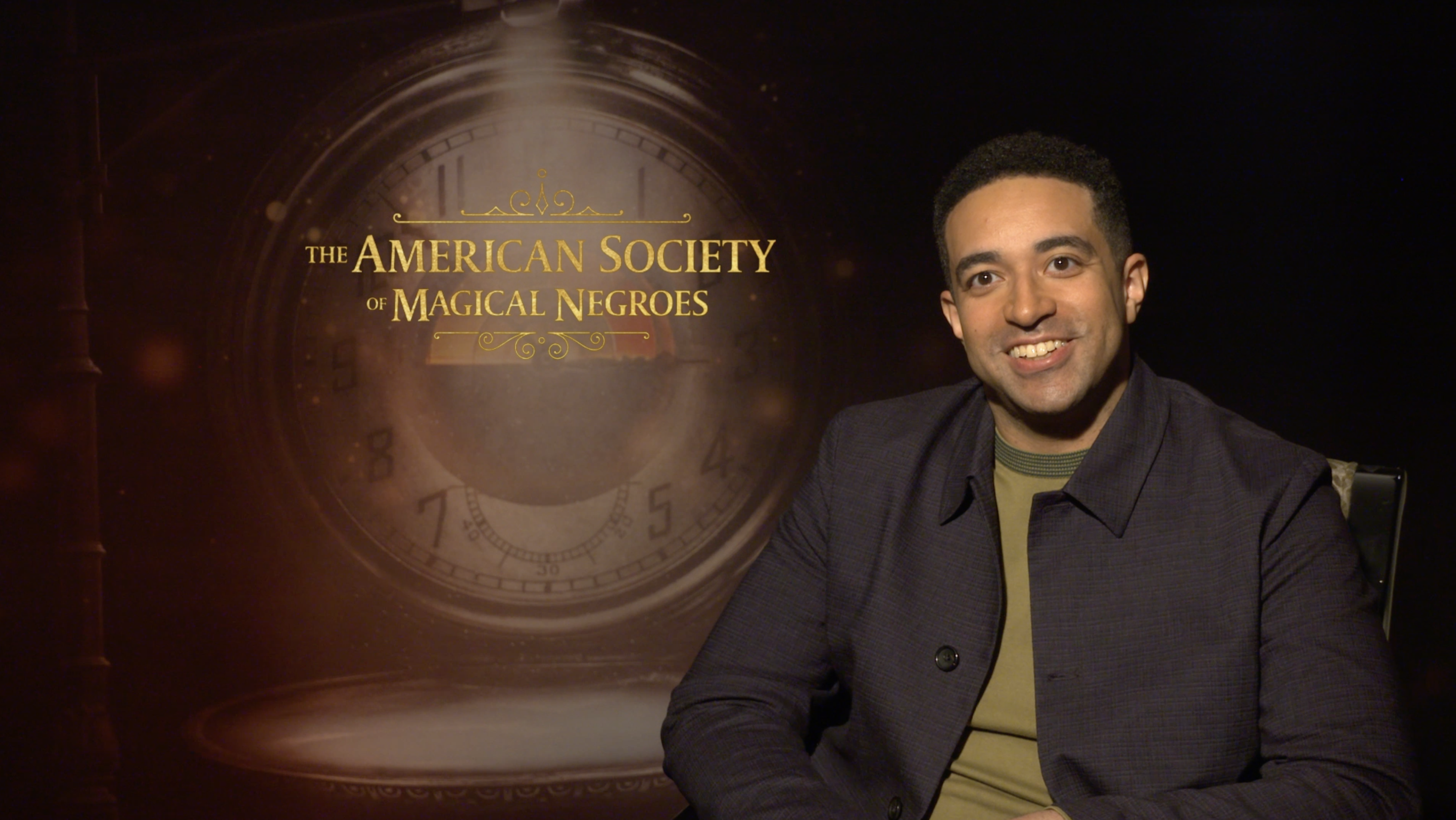 Kobi Labii smiles at the camera against a 'American Society for Magical Negroes' background