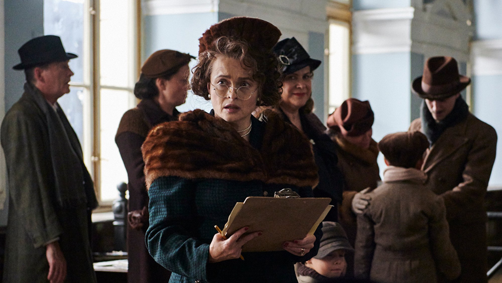 A woman wearing glasses stands holding a clipboard while other men and women mill around behind her.