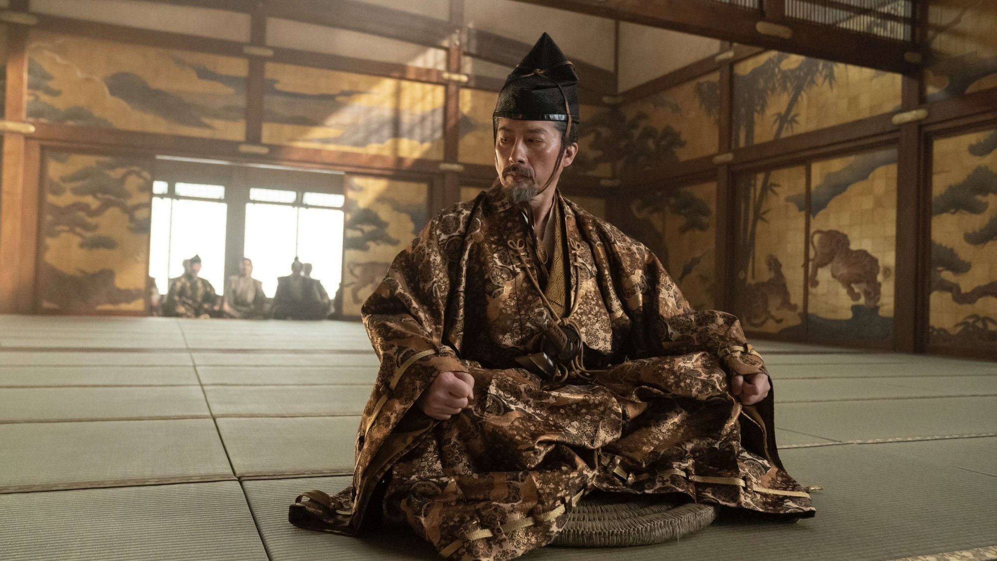 Yoshii Toranaga sits in a castle hall in an elaborate gold outfit.