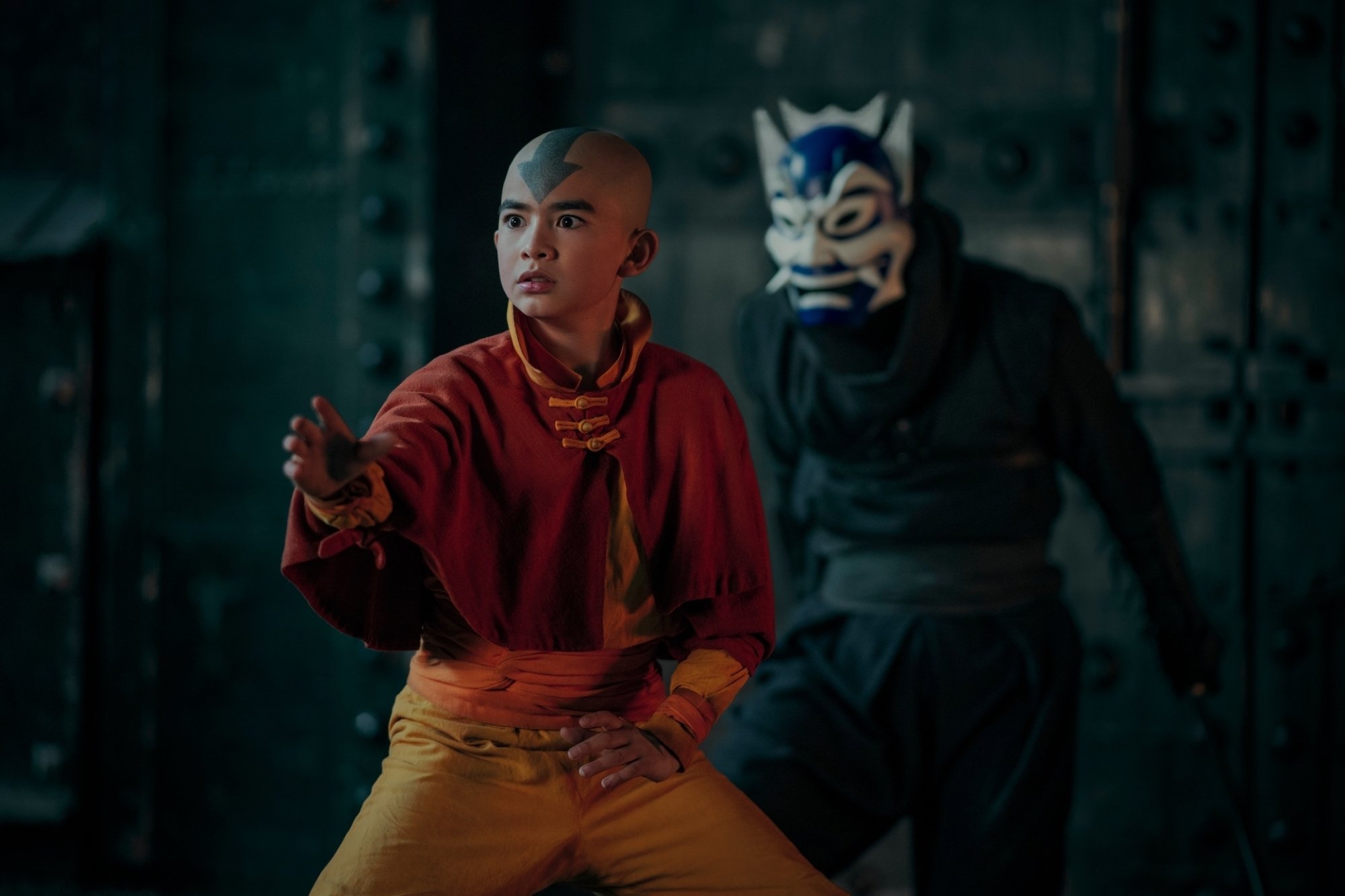 Aang stands in the foreground, as someone in a blue mask clad in black stands behind him.