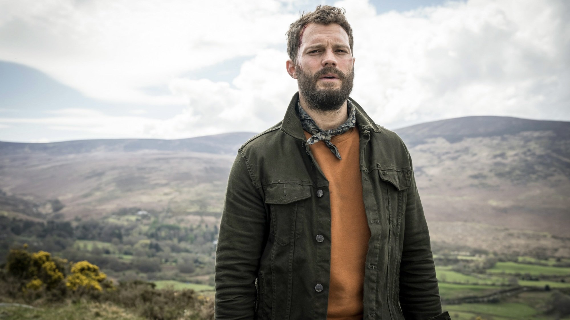 A man in a green jacket in the middle of a green, hilly landscape.