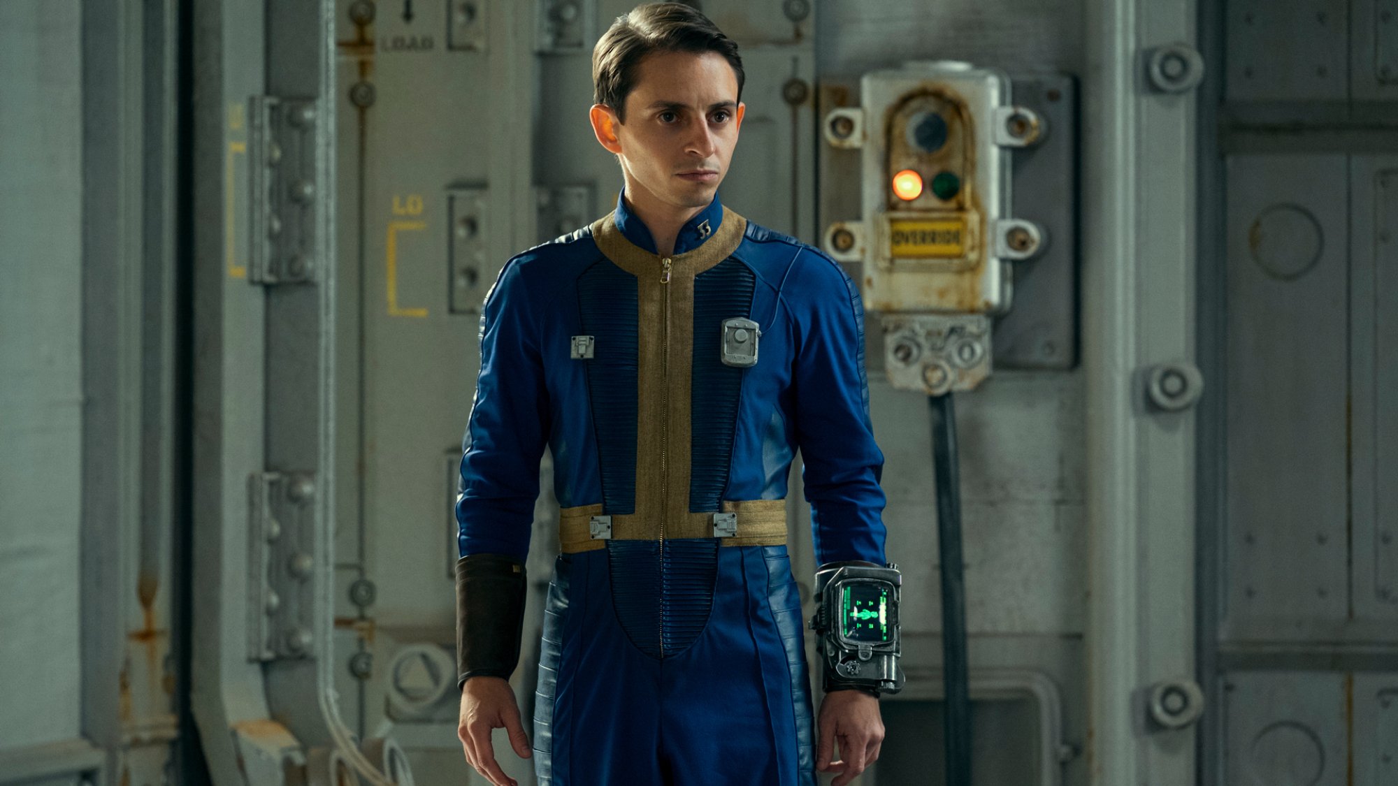 Moisés Arias wears a blue and yellow jumpsuit and a Pip-Boy on his wrist in "Fallout."